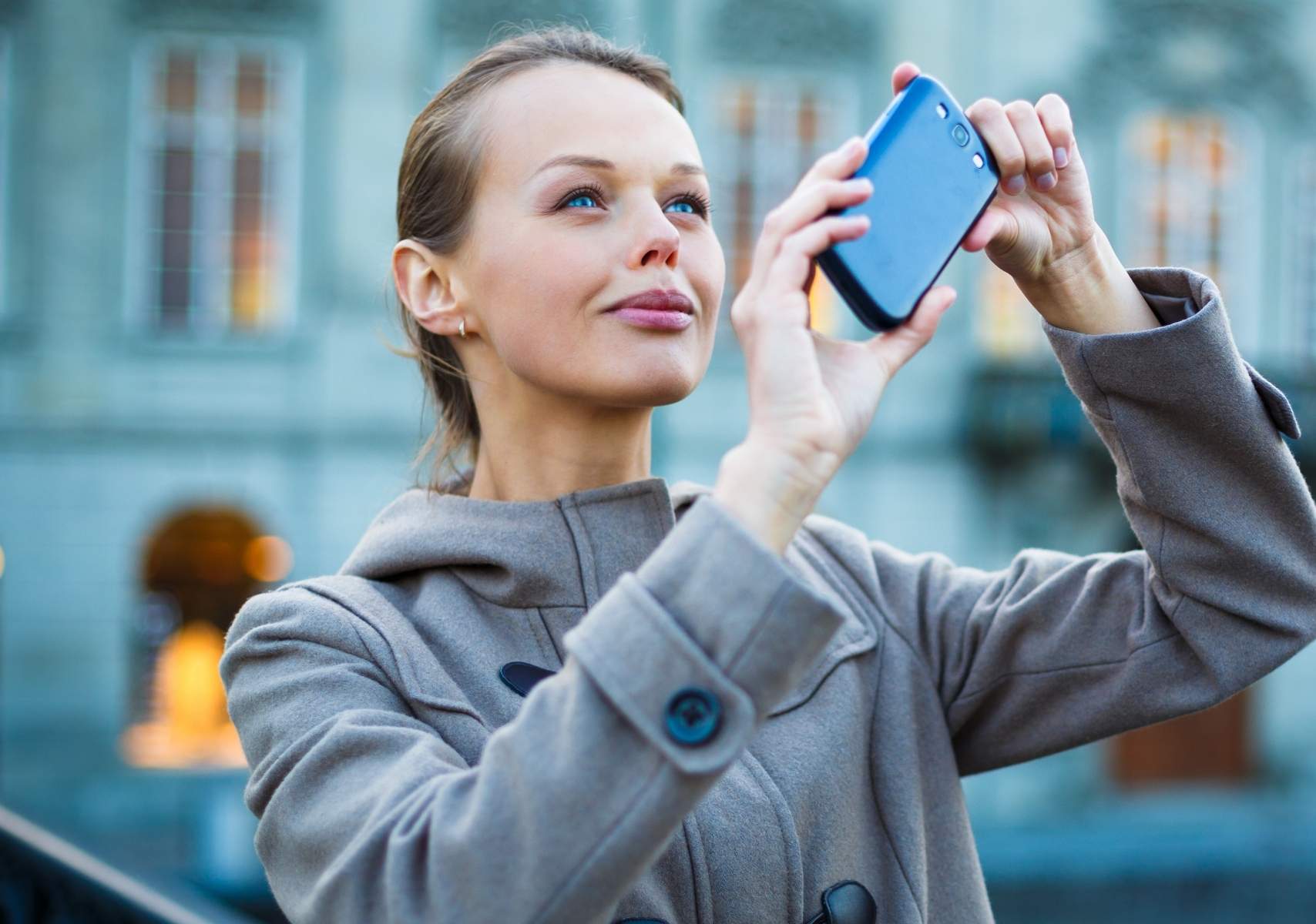 Mujer rubia de ojos azules, vestida con un abrigo gris, tomando una foto con celulares azul oscuro, mirando hacia arriba con expresión concentrada. Fondo urbano desenfocado.