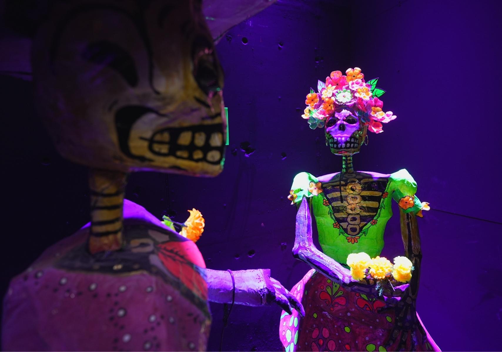 Catrinas de Día de muertos con un fondo obscuro y luces moradas en la experiencia Odisea México