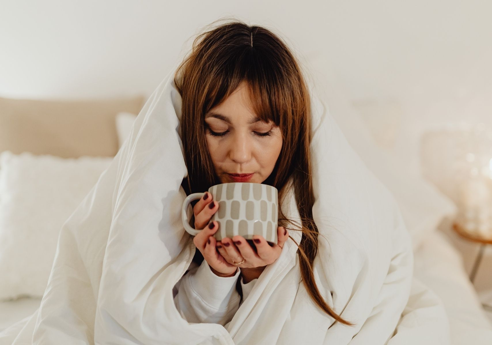 Mujer envuelta en una manta blanca y tomando cafe de una traza que sostiene con las dos manos en el invierno 2025